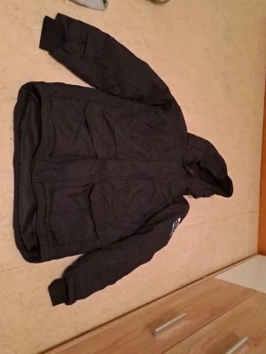 Svart vinterjacka North Pole Adventure - Jag säljer en svart vinterjacka från North Pole Adventure med huva och stora fickor framtill. Jackan har quiltad design och ribbade muddar vid ärmsluten. Perfekt för kalla dagar och riktigt skön att bära. Broderad patch på ärmen för extra stil. I bra skick. Säljs för 500 kr. 