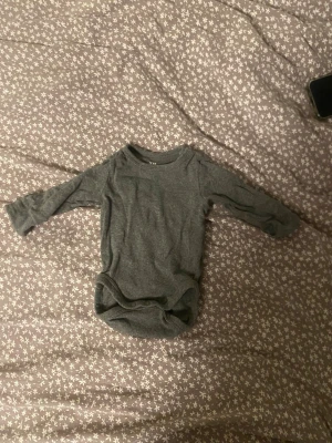 Grå basic body från H&M, stl 62 - Mjuk och enkel grå långärmad body från H&M i ekologisk bomull. Perfekt basplagg med rund hals och tryckknappar i grenen för smidig påklädning. Passar till allt och är superbekväm för de minsta.