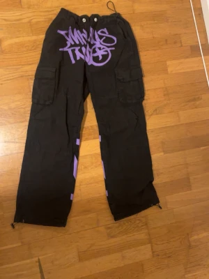 Svarta cargopants med lila tryck - Säljer ett par svarta cargopants med stora lila graffiti-tryck både fram och bak. Byxorna har flera fickor på sidorna, dragsko i midjan och vid bensluten samt coola detaljer längs benen. Perfekta för dig som vill sticka ut med streetwear-vibe.