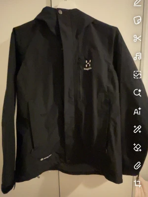 Svart vindjacka från Haglöfs - Säljer en svart vindjacka från Haglöfs med huva och dragkedja framtill. Jackan har en bröstficka med dragkedja och är tillverkad i slitstarkt material från Gore-Tex. Perfekt för blåsiga dagar och outdoor-äventyr.