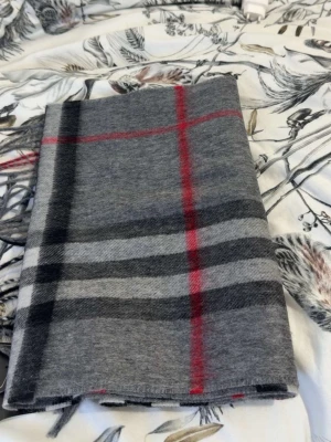 Burberry  halsduk  - Säljer en snygg burberry halsduk med klassiskt rutigt mönster i svart, vitt och rött. Halsduken har fransar i ändarna och är mjuk och varm, perfekt för kyliga dagar. Passar till de flesta outfits och ger en stilren touch.