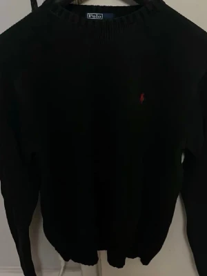 Svart stickad tröja Polo Ralph Lauren - Klassisk svart stickad tröja från Polo Ralph Lauren med rund hals och röd broderad logga på bröstet. Tröjan har lång ärm och ribbade muddar vid ärmslut och nederkant. Perfekt till lager på lager under en sen sommarkväll. Kom privat vid funderingar!