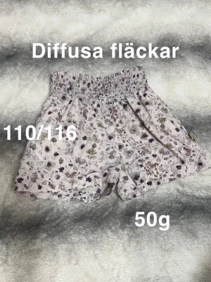 Blommiga shorts med resårmidja - Säljer ett par ljusa shorts med blommigt mönster och bred resår i midjan. Perfekta för varma dagar och superbekväma tack vare den lösa passformen. Materialet känns mjukt och svalt. Snygga att matcha med en enkel t-shirt!🌸 Diffusa fläckar. Storlek 110/115