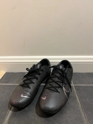 Svarta Nike fotbollsskor - Säljer ett par svarta Nike fotbollsskor med snyggt mönstrad ovandel och klassisk Nike-logga i silver. Skorna har snörning och är tillverkade i syntetmaterial för bra grepp och känsla på planen. Perfekta för dig som vill ha stilrena och funktionella skor till matchen.
