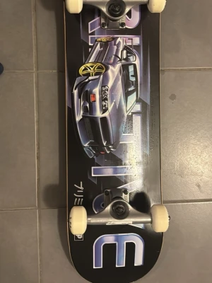 Primitive RPM svart skateboard 8.25" - Säljer en Primitive RPM komplett skateboard i storlek 8.25". Brädan har svart botten med en stor grafik av en sportbil i silver och lila toner samt texten PRIMITIVE. Hjul och trucks är vita och silverfärgade. Perfekt för dig som gillar street och snygg design.