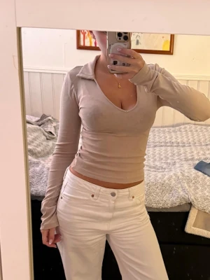 Beige långärmad topp - Säljer en beige långärmad topp från Gina Tricot i storlek XS. Toppen är figurnära med v-ringning och krage, samt croppad passform. Perfekt att styla med jeans eller kjol för en snygg look.