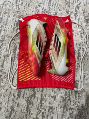 Adidas F50 fotbollsskor röd/vit - Säljer ett par Adidas F50 fotbollsskor med snygg design i rött, gult och svart. Skorna har snörning, platt sula med dobbar och är tillverkade i syntetmaterial. Medföljer röda innersulor och en matchande röd påse. Perfekt för dig som vill sticka ut på planen! Det finns en hål på toppen av skorna men inget annat. De går enkelt att fixa. Kan diskutera priset och de kommer med allt priset inte inte i hugget.