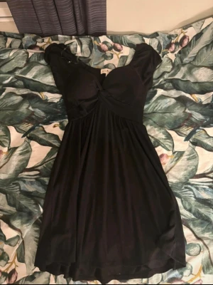 Svart kortklänning från Derek Heart - Supersnygg svart kortklänning från Derek Heart med ribbad struktur och twistad detalj framtill. Klänningen har korta ärmar och markerad midja som ger en smickrande siluett. Perfekt för dig som gillar enkel men chic stil.