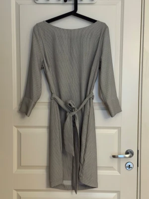 Klänning  - klänning med knytband i midjan. Klänningen har trekvartsärm med bred manschett och knappdetalj. Materialet är kavajliknande. 