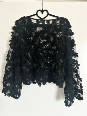 Svart blommig meshblus med volang - Säljer en svart transparent meshblus med 3D-blommor över hela tyget. Blusen har långa ärmar och en croppad passform. Perfekt för dig som vill sticka ut med en unik och trendig topp.