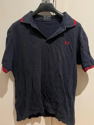 Marinblå Fred Perry pikétröja - Klassisk marinblå pikétröja från Fred Perry med röd broderad logga på bröstet och röda ränder på krage och ärmslut. Tillverkad i 100% bomull och har korta ärmar samt en avslappnad passform. Perfekt för en sportig och stilren look.