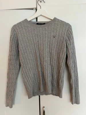 Grå kabelstickad tröja från GANT - Säljer en grå kabelstickad tröja från GANT i stretchig bomull. Klassisk rund halsringning och diskret logga på bröstet. Perfekt till jeans eller chinos för en clean look. Långa ärmar och ribbade muddar för extra komfort.