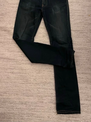 Mörkblå jeans från Nudie Jeans - Säljer ett par mörkblå jeans från Nudie Jeans med klassisk femficksdesign och kontrastsömmar. Jeansen har raka ben och en snygg tvätt som ger en schysst vintagekänsla. Märkespatch bak i midjan och diskret Nudie-logga vid bakfickan.