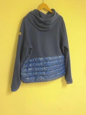 Blå hybrid pufferjacka från Moncler - Säljer en unik blå hybridjacka från Moncler med huva och quiltad pufferdetalj i blankt material nedtill. Jackan har långa ärmar, knapp vid halsen och dragsko i nederkant. Perfekt för dig som vill ha en chill och snygg look. Storlek L men passar M