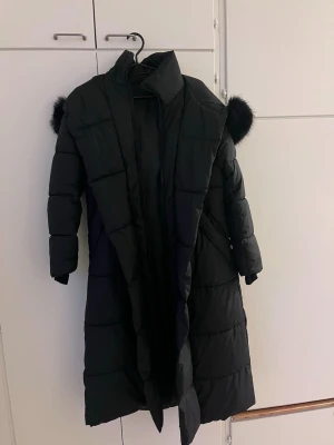 Svart lång dunjacka med päls - Säljer en svart lång pufferjacka med huva och päls. Jackan har quiltad design, dragkedja framtill och långa ärmar. Perfekt för kalla dagar och riktigt snygg till vintern. strl S! 
