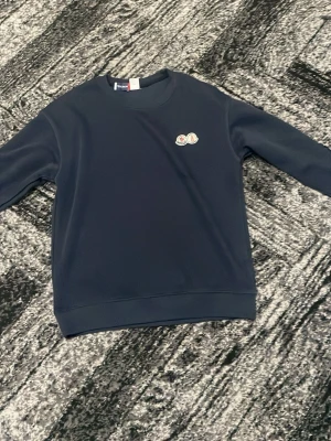 Mörkblå sweatshirt från Moncler - Säljer en stilren mörkblå sweatshirt från Moncler med broderade loggor på bröstet och ärmen. Tröjan har rund halsringning och långa ärmar. Perfekt för dig som gillar exklusiva streetwear-plagg.