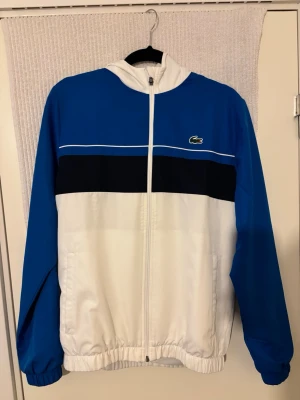 Blå och vit vindjacka från Lacoste - Säljer en snygg vindjacka från Lacoste Sport i blått, vitt och svart.  Helt ny har haft på den 2-3 gånger 