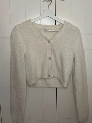 Vit croppad kofta från Lindex - Mysig vit croppad kofta från Lindex med fluffig känsla och glittriga knappar framtill. Koftan har lång ärm, v-ringning och ribbade muddar. Perfekt för dig som vill ha en enkel men ändå extra detalj i garderoben.