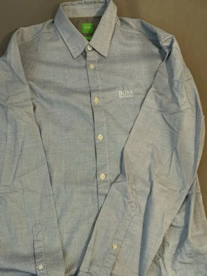 Blå skjorta från Hugo Boss - Snygg blå skjorta från Hugo Boss med diskret rutigt mönster och broderad logga på bröstet. Regular fit med klassisk krage, långa ärmar och vita knappar. Perfekt för dig som vill ha en stilren och clean look.