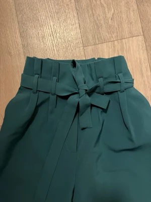 Mörkgröna byxor med knyt från BikBok - Snygga mörkgröna byxor från BikBok med hög midja och ett brett knytband i midjan. Byxorna har raka ben och ett stilrent, lite loose fit. Materialet känns mjukt och har en lätt struktur, perfekt för en chill men ändå uppklädd look. Materiallet är väldigt lätt och kön. PRISET KAN DIKUTERAS 