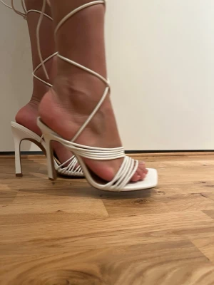 Vita sandaletter med snörning och klack - Säljer ett par trendiga vita sandaletter med höga klackar och tunna remmar som snöras runt vristen. Skorna har fyrkantig tå och öppen design, vilket ger en modern och stilren look. Tillverkade i syntetmaterial med brun innersula.