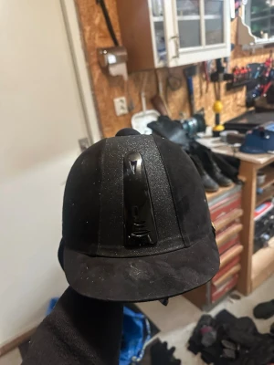 Svart ridväst från Kylin, strl 56 - Säljer en svart ridväst från Kylin med snyggt glittrigt tyg på ovansidan och diskreta ventilationshål på sidorna. Västen har en klassisk form och är tillverkad i ett mjukt syntetmaterial med mockakänsla. Perfekt för dig som vill ha stil på ridbanan.