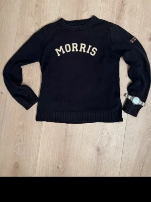 stickad tröja från Morris -  stickad tröja från Morris med krämfärgad logga broderad över bröstet. Klassisk rund halsringning, ribbade muddar och nederkant. Union Jack-detalj på ena ärmen. Perfekt till jeans för en clean och stilren look.