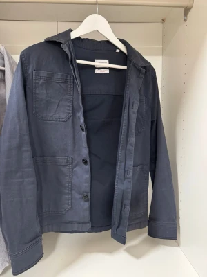 Mörkblå overshirt från Lindbergh - Säljer en mörkblå overshirt från Lindbergh med fyra stora fickor framtill och knäppning med svarta knappar. Jackan har klassisk krage och är tillverkad i bomull, perfekt för lager på lager. Enkel och stilren design som funkar till det mesta. Storlek xs passar som S. Hör av er med frågor