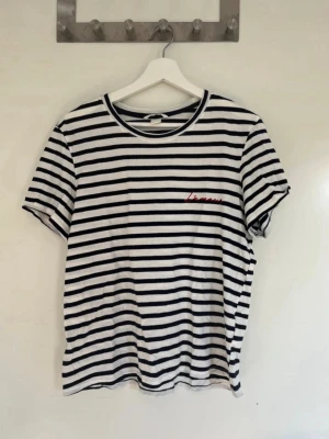 Randig t-shirt från H&M i bomull - Säljer en randig t-shirt från H&M i vitt och marinblått. T-shirten har korta ärmar, rund halsringning och ett litet rött broderi på bröstet. Tillverkad i mjuk bomull.