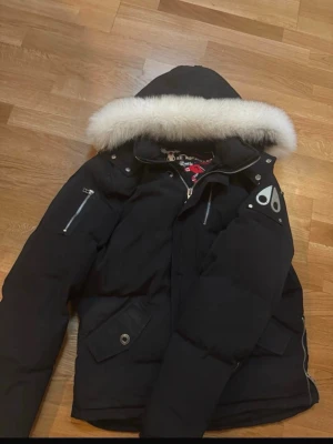 Svart Moose Knuckles dunjacka M - Säljer en svart dunjacka från Moose Knuckles med stor luva och vit fuskpäls. Jackan har flera dragkedjor, metallogo på ärmen och broderad insida med hockeymotiv. Perfekt för kalla dagar och riktigt snygg streetstil. De finns ba de såra felet med zippen längst nere men den funkar endån o zippa upp 