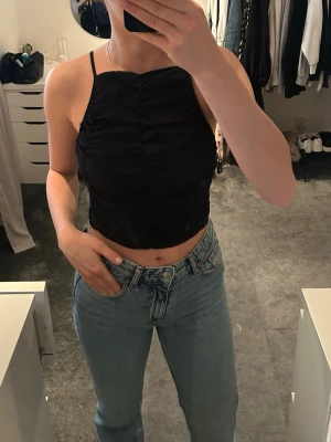 Svart croppad linnetopp med smala band - Säljer en svart croppad linnetopp med smala axelband och rynkade detaljer framtill. Toppen har en rak halsringning och sitter tight mot kroppen. Perfekt till sommaren 