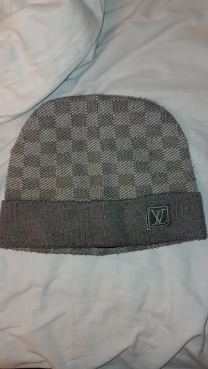  rutig mössa - Snygg grå mössa  med klassiskt rutigt mönster och uppvikt kant. Logga broderad framtill ger en exklusiv vibe. Stickad i mjukt material som håller dig varm och stilren under kyliga dagar.