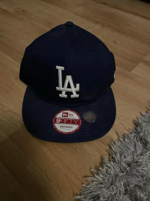 Mörkblå LA New Era 9Fifty keps - Säljer en mörkblå snapback-keps från New Era med LA-logga framtill. Modellen är 9Fifty och kepsen är tillverkad i 100% bomull. Klassisk rak skärm och justerbar baktill. Perfekt för dig som gillar sportig stil.