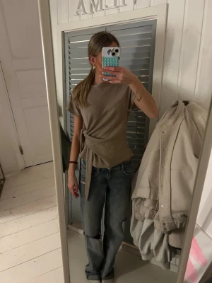 Beige topp med knyt från Zara - Säljer en beige topp från Zara i storlek S. Toppen har korta ärmar, rund halsringning och en snygg knytning i sidan som ger en cool och modern look. Materialet är mjukt och känns skönt mot huden. Perfekt att styla med jeans eller kjol.