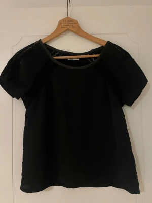 Ida Sjöstedt blus med spetsdetaljer - Säljer en svart blus med korta ärmar och fina spetsdetaljer längs axlar och ärmslut. Blusen har en rund halsringning och är gjord i ett lätt, tunt material som ger ett luftigt intryck. Perfekt för dig som gillar stilrena plagg med en twist.