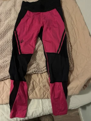 Träningstights från Craft i rosa och svart - Snygga träningstights från Craft i storlek S. Tightsen är rosa och svarta med coola paneler och Craft-logga på benet. Tillverkade i stretchigt material som består av återvunnen polyamid och elastan, perfekta för träning och sport.