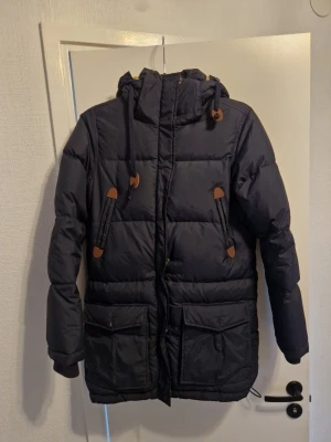 Mörkblå pufferjacka Race Marine - Säljer en mörkblå pufferjacka från Race Marine med huva och snygga bruna detaljer. Jackan har stora fickor framtill, dragkedja och snörning i huvan. Perfekt för kalla dagar när du vill hålla dig varm och stilsäker.