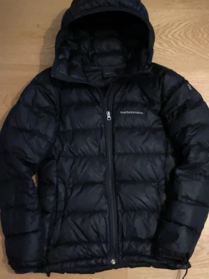 Svart dunjacka Peak Performance - Säljer en svart dunjacka från Peak Performance med huva och dragkedja framtill. Jackan är quiltad och har en clean look med logga på bröstet och ärmen. Perfekt för kalla dagar och riktigt skön att bära.