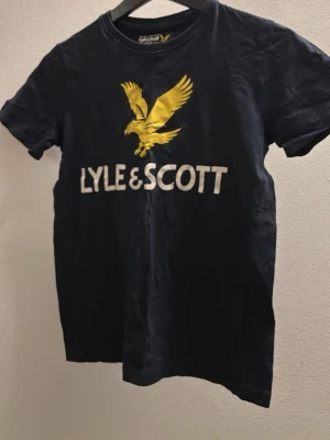 Marinblå Lyle & Scott t-shirt - Snygg marinblå t-shirt från Lyle & Scott med stort gult örntryck och vit logga på bröstet. Klassisk rund hals och korta ärmar, perfekt till jeans eller shorts. Skön och mjuk bomullskvalitet.