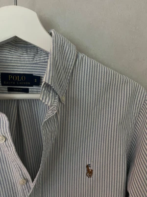 Ralph Lauren Skjorta - Riktigt stilig Ralph Lauren skjorta i dunder skick och randig med vit/ljusblå färg. Skjortan är i storlek S och den mest eftertraktade modellen Slim Fit. Vid frågor eller funderingar tveka inte på att höra av er! 😊