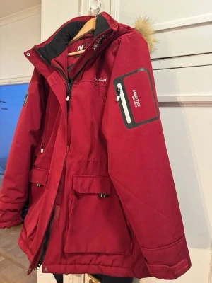 Röd parkas från True North XL - Säljer en röd parkas från True North i storlek XL. Jackan har flera praktiska fickor, dragkedja, huva med fuskpäls och detaljer som patch på ärmen. Materialet är tåligt och fodrat, perfekt för kalla dagar. Använt få gånger 