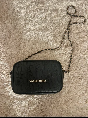 Svart axelväska från Valentino - Säljer en svart axelväska från Valentino med präglad logotyp över hela väskan. Väskan har en kedja med svarta och guldfärgade detaljer samt guldfärgad dragkedja med Valentino-gravyr. Stilren och kompakt design, perfekt för att bära det viktigaste.