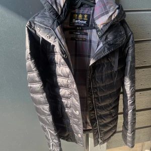 Svart quiltad jacka från Barbour - Säljer en svart quiltad jacka från Barbour med huva och dragkedja. Jackan har ett klassiskt rutigt innerfoder och stängs med både dragkedja och knappar. Perfekt för kyligare dagar och snygg till många outfits.