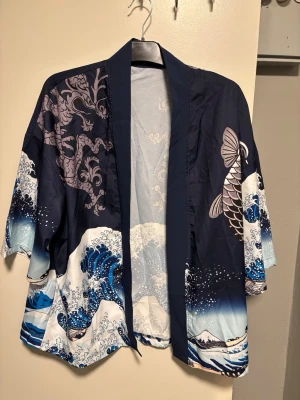 Blå kimono-skjorta med drakmotiv - Säljer en riktigt snygg kimono-inspirerad skjorta med korta ärmar och öppet frontparti. Skjortan har ett fett tryck med vågor, drake och koi-fisk i blått, vitt och lila. Materialet känns lätt och luftigt, perfekt för chill dagar. Unik design som sticker ut i mängden.