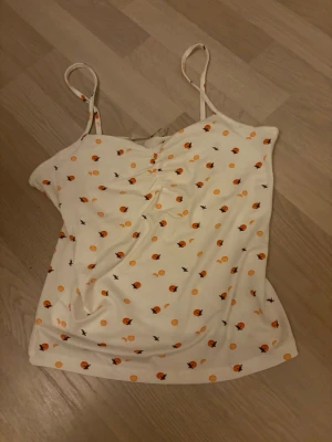 Vit linnetopp med apelsinmönster - Supersöt vit linnetopp med tunna axelband och tryck av små apelsiner och apelsinskivor i orange. Toppen har lätt rynk framtill och är perfekt för varma dagar. Materialet känns mjukt och svalt mot huden. Oanvänd 