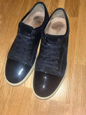 Mörkblå sneakers från Lanvin - Snygga mörkblå sneakers från Lanvin med ovandel i mocka och lackad tå. Skorna har vita sulor, rund tå och klassisk snörning. Perfekta för dig som vill ha en stilren men ändå unik sneaker med lyxig känsla. Byten accepteras 