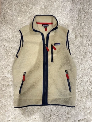 Beige fleeceväst från Patagonia - Säljer en beige fleeceväst från Patagonia med marinblå detaljer och röda dragkedjor. Västen har hög krage, tre fickor med dragkedja och ett klassiskt Patagonia-märke på bröstet. Perfekt att slänga på sig när det är lite kyligt ute.