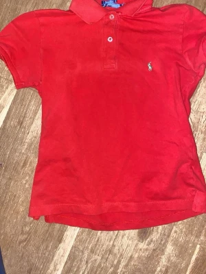 Röd pikétröja från Polo Ralph Lauren - Snygg röd pikétröja från Polo Ralph Lauren med klassisk krage och två knappar framtill. Tröjan har korta ärmar och den ikoniska broderade loggan på bröstet. Perfekt för dig som gillar stilrena och sportiga plagg.