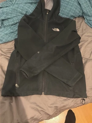 Svart fleecejacka från The North Face. Säljer då den ej passar mig längre. Väldigt skön  - Svart fleecejacka med huva från The North Face. Jackan har dragkedja framtill, två sidofickor med dragkedja och broderad logga på bröstet. Mjuk insida och klassisk passform, perfekt för kyliga dagar.