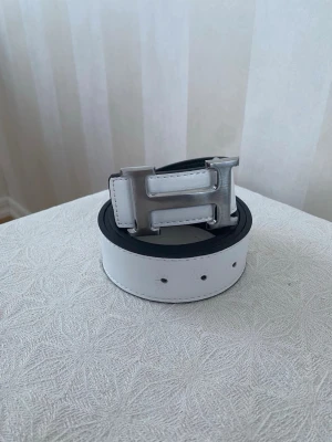 Hermes bälte vit - Säljer ett vändbart bälte i vitt och svart skinn med ett stort H-format metallspänne. Bältet har stilrena sömmar och en lyxig känsla tack vare det blanka spännet. Perfekt för att lyfta din outfit med en snygg detalj.